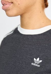 Zwarte T-shirt met korte mouwen en een witte geribbelde kraag; heeft een klein wit Adidas-logo op de voorkant. Gladde textuur en casual ontwerp.