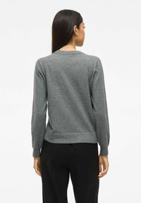 Pull gris à manches longues, col rond et texture douce. L'ourlet est légèrement plus court à l'arrière. Porté avec un pantalon noir.