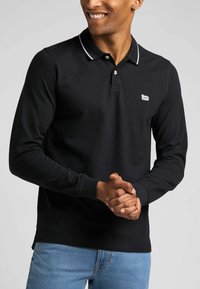 Zwarte langslopen polo met witte kraagtrim en een klein geborduurd logo op de borst, gemaakt van zacht stof, in combinatie met lichtblauwe jeans.