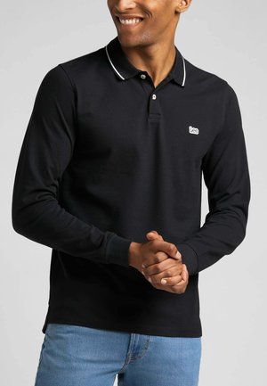 Zwarte langslopen polo met witte kraagtrim en een klein geborduurd logo op de borst, gemaakt van zacht stof, in combinatie met lichtblauwe jeans.