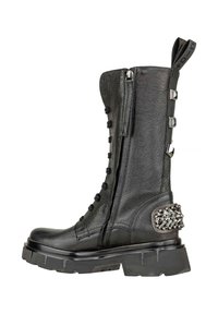 Stivale in pelle nera fino al polpaccio con cerniera laterale, lacci anteriori, suola spessa, borchie decorative sul tallone e un tassello per il sollevamento in cima.