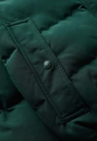 Fragment ciemnozielonej kurtki puchowej z widocznymi przeszyciami, gładką teksturą oraz detalem zatrzasku na naszywanej kieszeni.