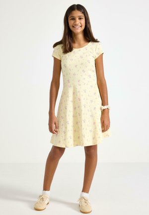 Hymyilevä tyttö seisoo, hänellä on lyhythihainen keltainen mekko, jossa on pieniä kukkakuoseja, valkoiset nilkkasukat, beiget lenkkarit ja rannassaan hiusdonitsi.