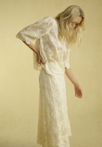 Blusa in pizzo con maniche a tre quarti, di colore crema, abbinata a una lunga gonna coordinata. Dettagli di ricami floreali su tutta la superficie.