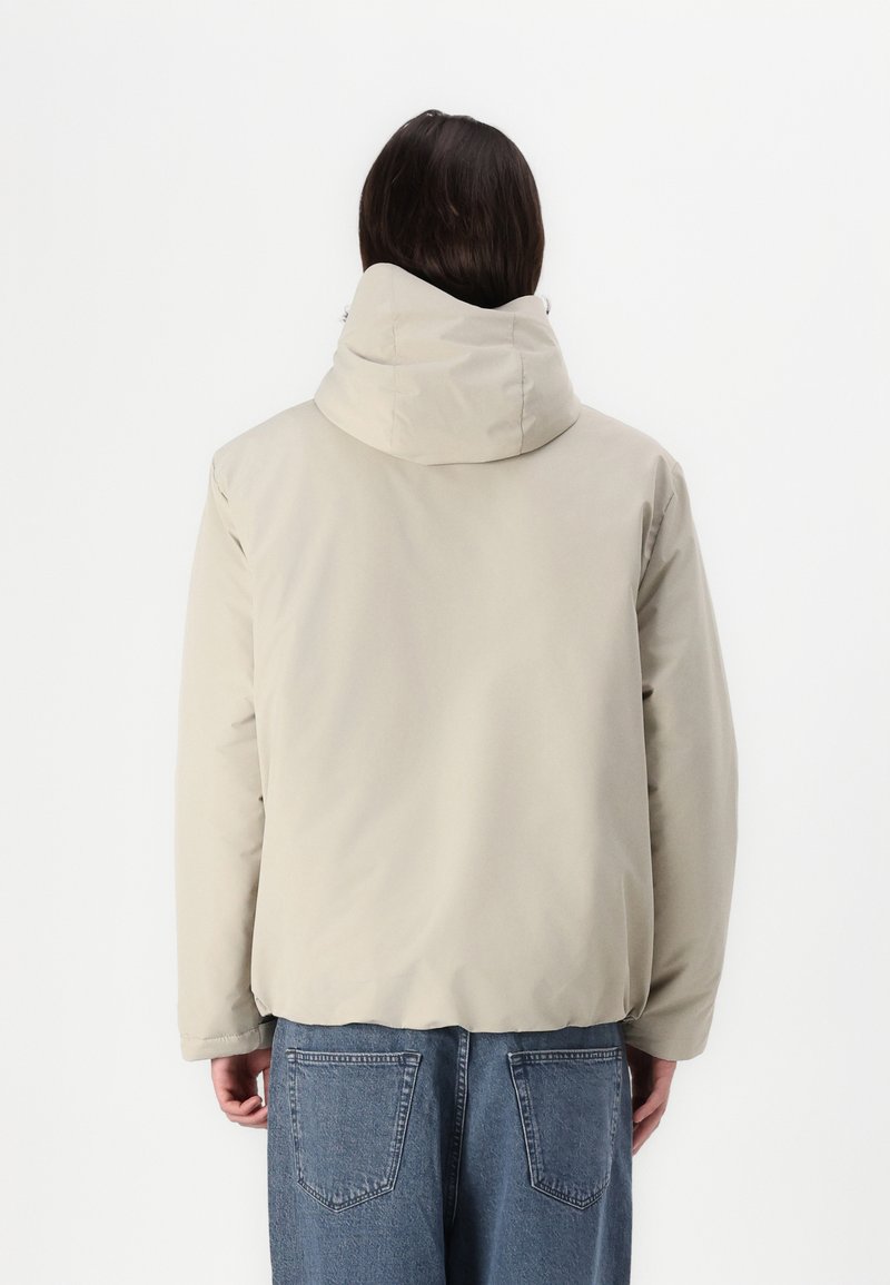 Chaqueta de plumas beige con un alto cuello, mangas largas y puños elásticos; presenta una textura suave y un corte relajado. Lleva puesta unos vaqueros azules.