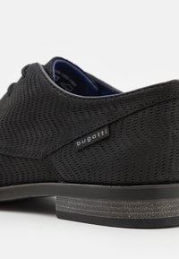 Chaussure noire avec une surface texturée présentant un motif en zigzag. Elle est fabriquée en matériau synthétique, avec un intérieur bleu contrastant et une étiquette de marque.