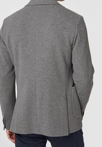 Blazer grigio realizzato in tessuto morbido, con una vestibilità sartoriale, rever con tacche e due tasche laterali con dettagli a bottone. Vista posteriore mostrata.