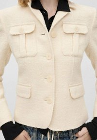 Beiger Blazer mit strukturierter Oberfläche und taillierter Passform, zwei Brusttaschen und drei Knöpfen vorne, kombiniert mit einem schwarzen Hemd darunter.