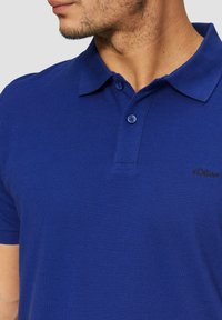 Niebieska koszulka polo z kołnierzykiem, krótkimi rękawami i teksturowanym materiałem. Posiada małe czarne logo na piersi. Luźny krój.