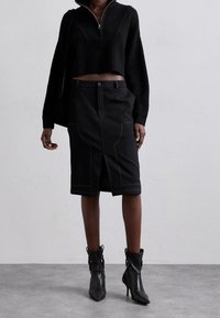 Pull noir court à col montant et texture côtelée, associé à une jupe crayon noire longueur genou avec surpiqûres contrastantes et fente à l'avant.