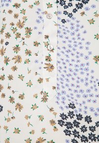 Camicia a bottoni floreale che presenta una base di tessuto bianco con fiori gialli, blu e neri, messa in risalto da un'apertura con bottoni perlati.