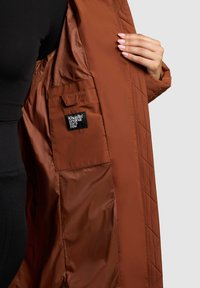 Braune gesteppte Jacke mit glänzender Innenseite, Seitentasche und einem schwarzen Etikett mit weißem Text. Das Design ist geschmeidig und hat eine glatte Textur.