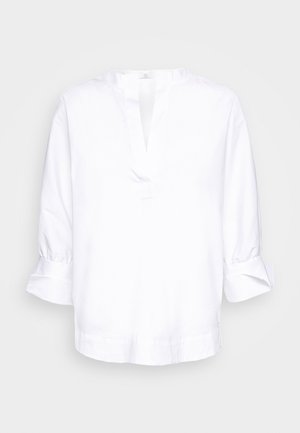 Blusa blanca de manga larga con abertura en V y puños doblados sobre fondo liso.