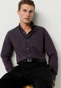 Dunkellila Hemd mit Knopfleiste, langen Ärmeln, spitzem Kragen und Brusttasche. Kombiniert mit schwarzen Hosen und einem schwarzen Gürtel.