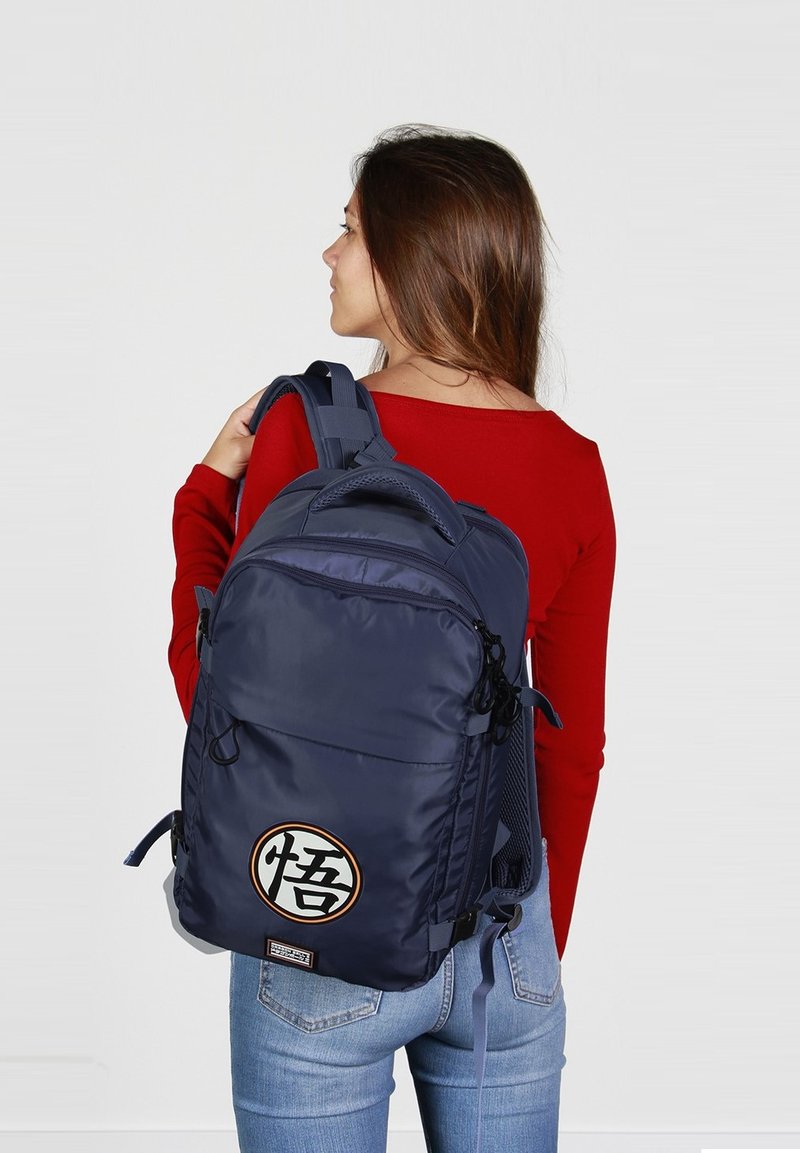 Sac à dos bleu marine avec un détail de logo rond, doté de bretelles réglables, de fermetures éclair et d'une finition texturée. Porté sur le dos d'une personne.