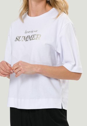 Femme portant un t-shirt blanc à manches courtes avec le texte "less monday more SUMMER" en lettres métalliques sur le devant.