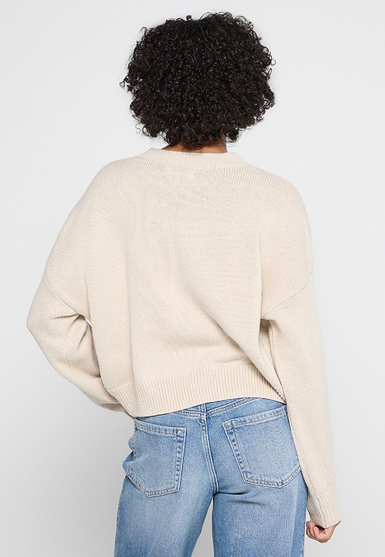 Maglione beige lavorato a maglia con una vestibilità rilassata, maniche ampie e orlo a coste. La trama appare morbida, progettata per il comfort e per essere indossata a strati.