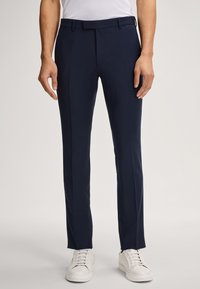 Navyblaue maßgeschneiderte Hose aus glattem Stoff, mit schlanker Passform, Gürtelschlaufen und gefaltetem Vorderteil, kombiniert mit weißen Sneakers.