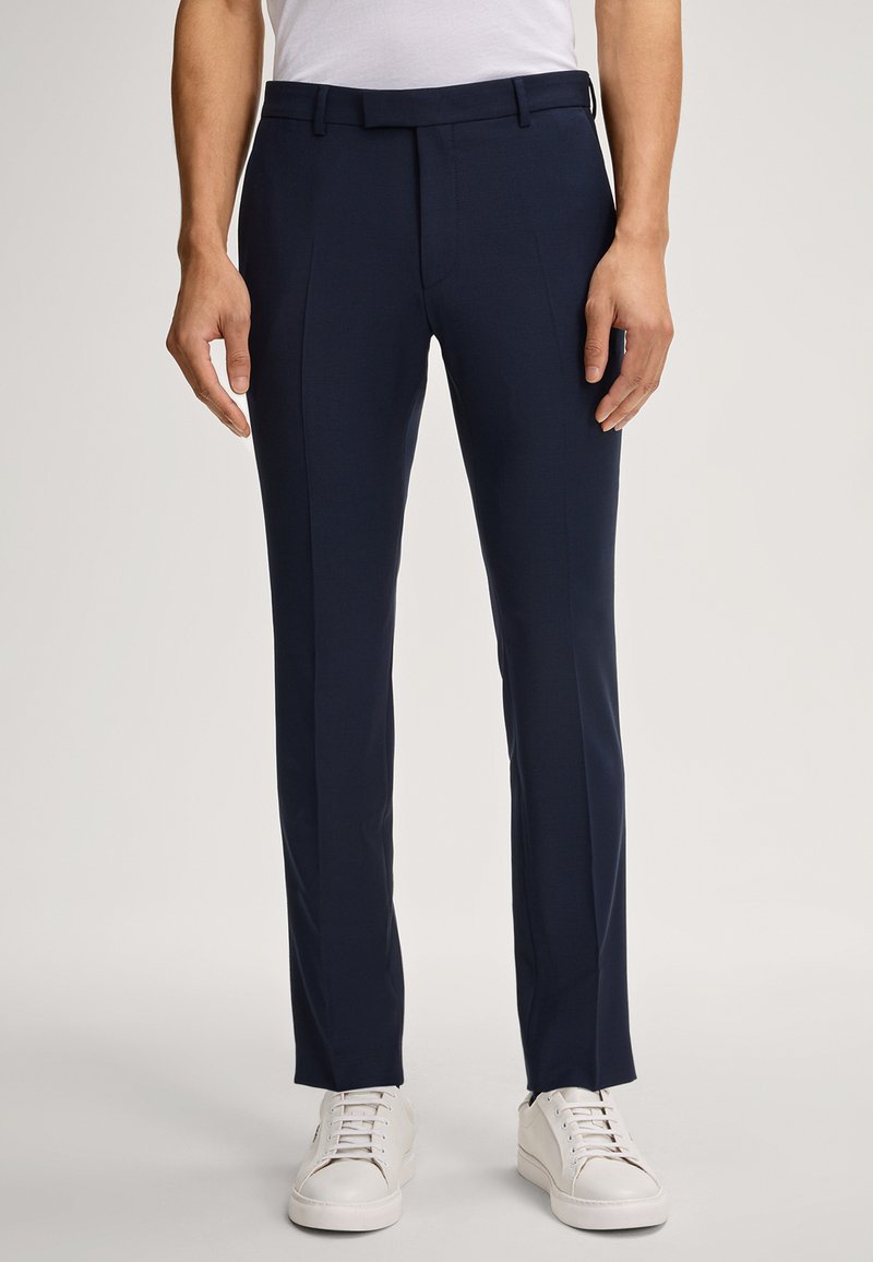 Navyblaue maßgeschneiderte Hose aus glattem Stoff, mit schlanker Passform, Gürtelschlaufen und gefaltetem Vorderteil, kombiniert mit weißen Sneakers.