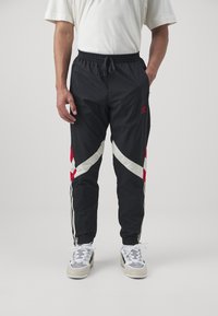 Černé sportovní kalhoty s červenými a bílými diagonálními akcenty, elastickým pasem, bočními kapsami a černými pruhy po délkách nohavic.