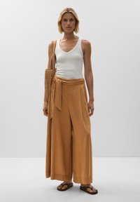 Lightweight, mustard-colored linen wide-leg pants with a tied waist and side pockets, paired with a white tank top and brown flip-flops.