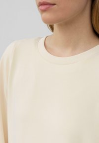 Maglia beige a maniche lunghe con scollatura rotonda e tessuto morbido. Texture liscia con una sottile cucitura attorno al colletto.
