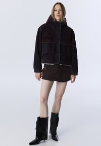 Tmavě modrá péřová bunda s kapucí, která má dvě náprsní kapsy a elastické manžety. Vytvořeno s hnědou mini sukní a černými kozačkami po kolena.