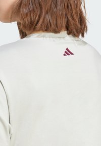 Bela bombažna majica s krožno ovratnikom, ki ima maroon Adidas logo na zgornjem delu hrbta. Gladka tekstura in sproščen dizajn.