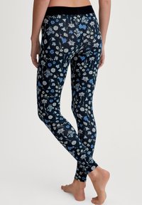 Persoon die blootvoets staat en zwarte leggings met blauw en witte bloemenpatronen draagt, gezien van achteren tegen een effen witte achtergrond.