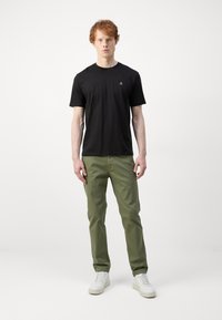 Zwart t-shirt met korte mouwen en een ronde hals, voorzien van een klein logo op de borst, gecombineerd met olijfgroene broek en witte sneakers.