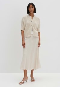 Cardigan beige en tricot à manches courtes, fermeture boutonnée, associé à une jupe longue couleur crème clair et des sandales à talons argentés.