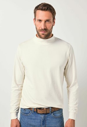Camisa de cuello alto de manga larga en color crema, hecha de una tela suave. Presenta mangas ajustadas, textura lisa y un diseño casual, combinada con unos vaqueros.