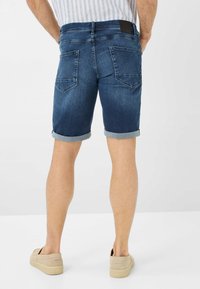 Shorts en denim bleu foncé avec un ourlet roulé, comprenant deux poches arrière et une étiquette noire sur la taille. Portés avec des chaussures à enfiler beige.