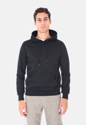 Uomo con capelli ricci corti, che indossa un hoodie nero e pantaloni cargo beige, in piedi davanti a uno sfondo bianco semplice.