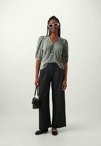 Blusa verde com padrão e mangas bufantes, combinada com calças largas pretas de cintura alta. Acessórios incluem uma pequena mala preta e óculos de sol.