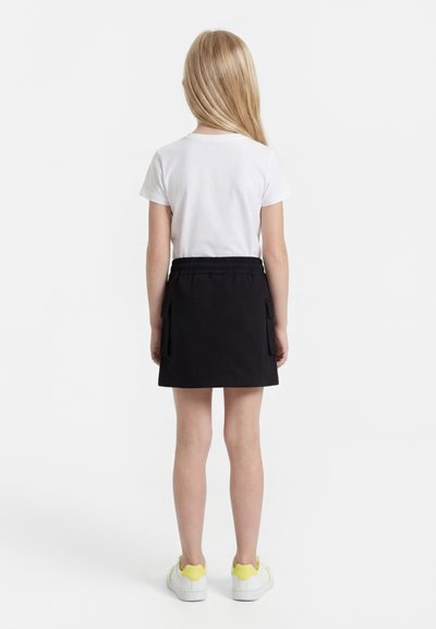 Blonde child standing facing away, wearing a white short-sleeve shirt, black skirt with cargo pockets, and white sneakers with yellow accents.