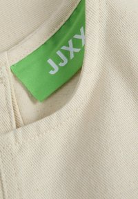 Tela de color crema con un acabado texturizado y costuras visibles. Etiqueta verde con "JJXX" en blanco, unida cerca del escote.