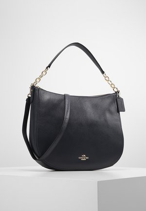 Černá kožená kabelka přes rameno se zlatými řetízkovými doplňky, zapínáním na zip a logem Coach na přední straně, vystavená na bílém podstavci.