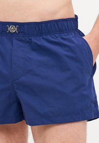 Pantaloncini da mare blu navy realizzati in tessuto leggero, con vita elastica, tasche laterali e un accento metallico decorativo sul davanti.