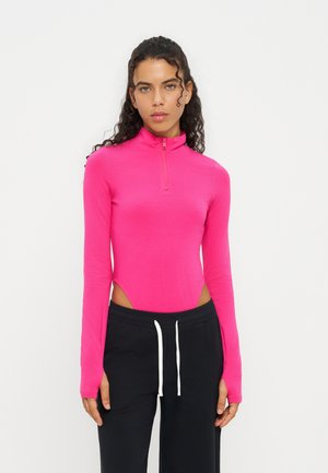 Femme aux cheveux bouclés et foncés portant un haut rose vif à manches longues avec fermeture éclair et un pantalon noir avec des cordons blancs.