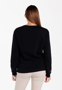 Sudadera negra de manga larga, con cuello redondo y puños acanalados. Tejido texturizado con detalle del logo en la manga. Combinada con pantalones de color claro.