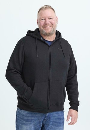 Lächelnder Mann mit kurzen Haaren und Bart, der einen schwarzen Zip-Hoodie und blaue Jeans trägt, und vor einem schlichten weißen Hintergrund steht.