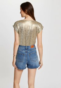 Kortærmet, metallisk guld top med afslappet pasform, parret med mid-rise, blå denim shorts med baglommer og et lædermærke.