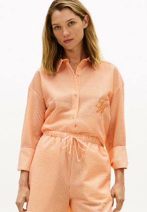 Mujer con camisa de botones de rayas naranja y blanca y pantalones a juego con cordón, contra un fondo blanco liso.