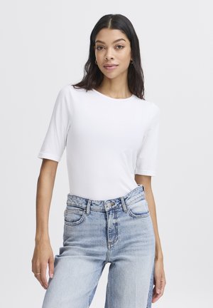 Femme aux cheveux foncés portant un t-shirt blanc uni à manches courtes, rentré dans un jean taille haute bleu clair, sur un fond uni.