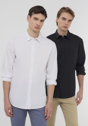 Due uomini stanno spalla a spalla. Uno indossa una camicia bianca con bottoni, l'altro una nera. Entrambe le camicie hanno maniche lunghe e un colletto classico.