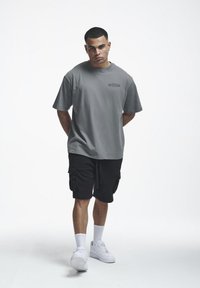 Jeune homme avec des boucles d'oreilles portant un t-shirt gris oversized, un short cargo noir, des chaussettes blanches et des baskets blanches, debout les mains dans le dos.