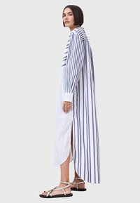 Lang gestreepte jurk in wit en blauw, met een hoge split, knoopmanchetten en een rechte kraag. Gemaakt met zilveren sandalen.