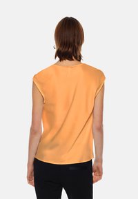 Top in seta arancione con maniche a cappuccio, caratterizzato da un colletto rotondo e una texture liscia, abbinato a pantaloni neri. Vista posteriore mostrata.