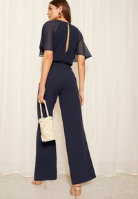 Marine jumpsuit met korte flapmouwen en een keyhole achterkant, voorzien van wijde pijpen. Gecombineerd met een gestructureerde crèmekleurige handtas.